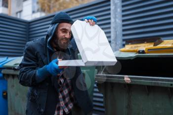 Bearded dirty beggar found pizza in trashcan on city street. Poverty is a social problem, homelessness and loneliness, alcoholism and drunk addiction, urban lonely