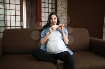 Pregnant woman with belly sitting on sofa and eating chocolate. Pregnancy, gluttony in prenatal period. Expectant mom resting at home