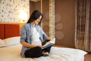 Pregnant woman with belly reading a book in bed at home. Pregnancy, calm in prenatal period. Expectant mom resting in bedroom