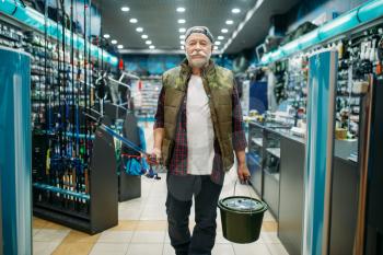 Fisherman with rod and plastic bucket in fishing shop. Male angler buying equipment and tools for fish catching and hunting, assortment on showcase in store