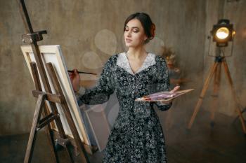 Female painter with brush and palette draws in art studio. Artist at her workplace, creative master at the easel in workshop