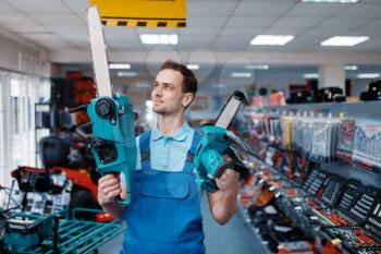 Male worker in uniform holds big and small chainsaws in tool store. Choice of professional equipment in hardware shop, instrument supermarket
