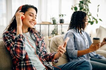 Two cute women in headphones leisures on sofa with cup of coffe. Pretty girlfriends in earphones relax in the room, sound lovers resting on couch, female person using mobile phone