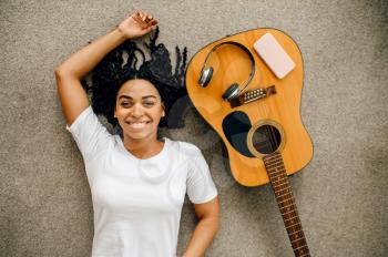 Cute woman with guitar and headphones lying on the floor at home, top view. Pretty lady with musical instrument relax in the room, female music lover resting