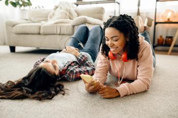 Laughing women in headphones enjoys listening to music lying on the floor at home. Pretty girlfriends in earphones relax in the room, sound lovers resting on couch, female friends leisures together
