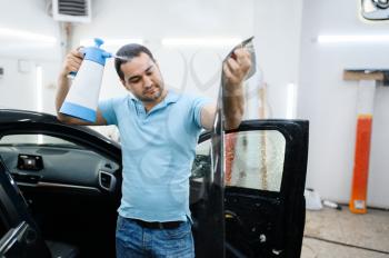 Male specialist with spray wetting car tinting, tuning service. Mechanic applying vinyl tint on vehicle window in garage, tinted automobile glass