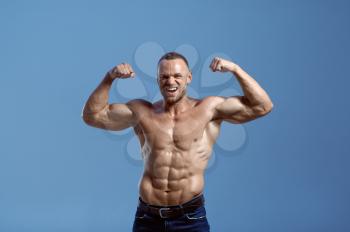Male athlete with muscular body shows his power in studio, blue background. One man with athletic build, shirtless sportsman in jeans pants, active healthy lifestyle