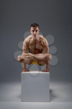 Male yoga keeps balanc on hands in difficult pose on the pedestal, meditation position, grey background. Strong man doing yogi exercise, asana training, top concentration, healthy lifestyle