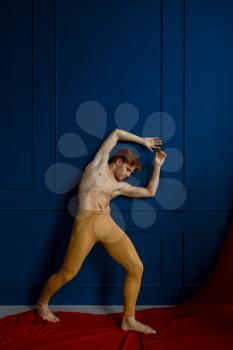 Male ballet dancer poses at blue wall in dancing class, red cloth on background. Performer with muscular body, grace and elegance of movements