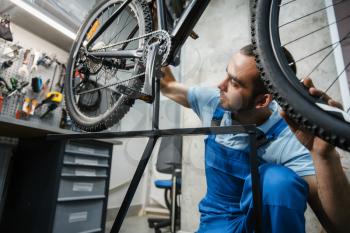 Bicycle repair in workshop, man checks the wheel for backlash. Mechanic in uniform fix problems with cycle, professional bike repairing service
