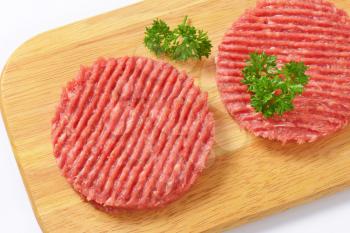 two raw hamburger patties with parsley on wooden cutting board