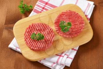 two raw hamburger patties with parsley on wooden cutting board and checkered dishtowel