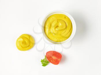 bowl and swirl of yellow mustard and slice of tomato on white background