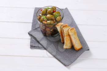 glass of marinated green olives with toast on grey place mat