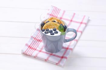 cup of american pancakes with white yogurt and fresh blueberries on checkered dishtowel