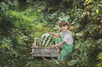A little girl carries a huge watermelon on a cart.
