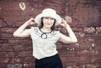 Pretty young woman in white blouse and hat posing against brick wall background. Woman holds the edges of the hat by hands. Toned photo with copy space. Vintage style photo.