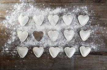 Raw ravioli in the shape of hearts, sprinkle with flour, on dark wooden background. Cooking dumplings. Top view.