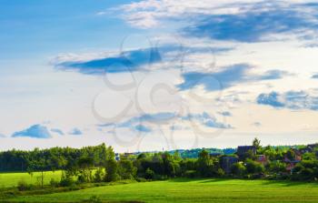 Sunny summer evening. Rural landscape. The sky with clouds, grass field, trees and farmhouses.