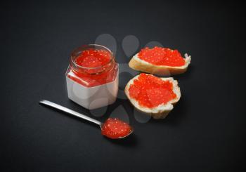 Fresh deliciousd caviar. Sandwiches with red caviar, glass jar and spoon on black background.