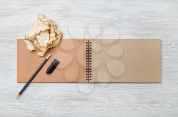Photo of open blank kraft notebook, pencil, eraser and crumpled paper on light wood table background. Responsive design template. Flat lay.