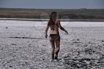 Woman walking along the dry bottom of a salt lake, rear view. Walk the dark-haired woman in a swimsuit on the bottom of a dry lake with salt and mud. The ancient dried-up lake.