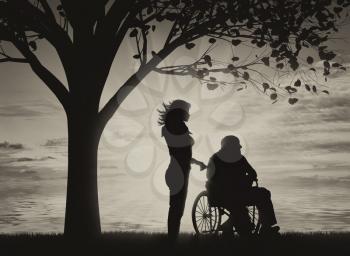 Silhouette of nurse caring for a disabled person in a wheelchair resting under a tree near sea. Concept of caring for a disabled person and house of aged