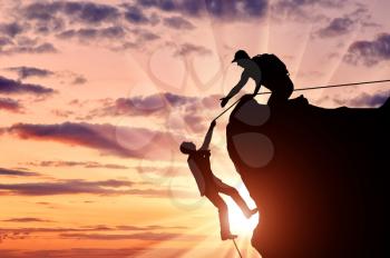 Silhouette of a climber who helps to climb the top of a man, throws him a rope and holds out his hand. Conceptual scene
