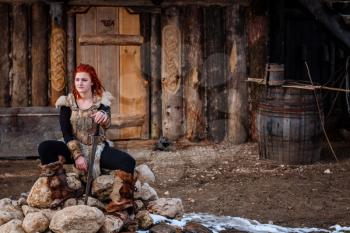 Red-haired woman is a Viking. Against the backdrop of a village or an ancient settlement. In the hands of a sword