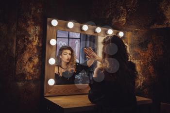 Beautiful fashion model woman posing near the mirror. Woman in a beauty salon looks at her reflection in the mirror with lamps and checks hairstyle and makeup