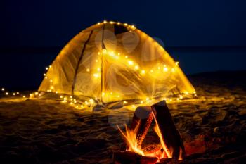The fire at night on the beach. Summer mood. Night camping with a retro garland on sea shore.