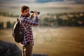 Guy looking at binoculars in hill. man in t-shirt with backpack. Young Caucasian man during hike in valley landscape