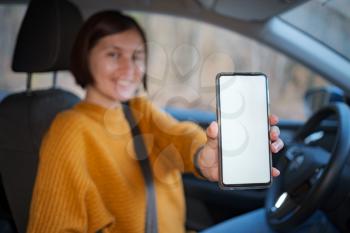 A young happy woman driving a car, she shows empty phone screen . Useful mobile apps for driver. Side view inside car, mockup