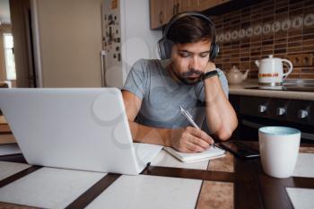 Focused young man wear headphones study online watching webinar podcast on laptop listening learning education course conference calling make notes sit at work desk, elearning concept