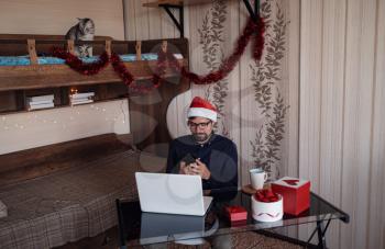 Happy male in deep blue shirt makes a purchase on the Internet via a phone. Lots of scenery for the new year. Working from home businessman wearing red Santa hat