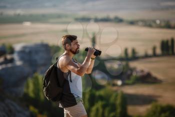 Guy looking at binoculars in hill. man in t-shirt with backpack. Young Caucasian man during hike in valley landscape