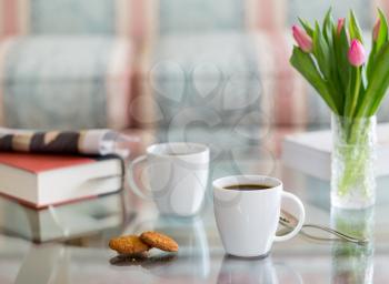 Modern white porcelain cup of black coffee on glass table with spoon and ginger biscuits. Book newspaper and vase of tulips in the background out of focus