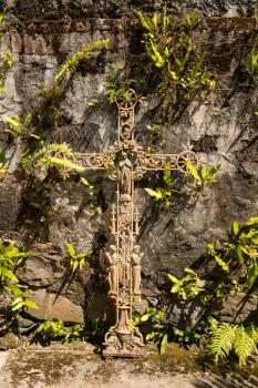 Ornate metal crucifix with statues of Mary and saints on old stone wall with plants growing into the shape