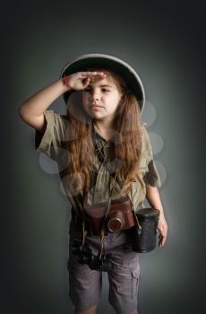 little girl in a tropical khaki uniform and a cork helmet with binoculars and a camera staring into the distance, covering herself with the palm of her hand