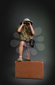 a little girl in a tropical khaki uniform and cork helmet looks through binoculars putting her foot on a suitcase