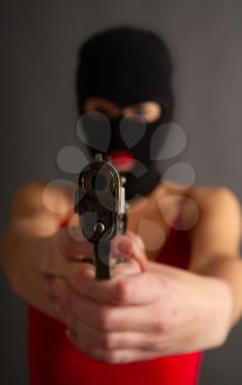 Young self-confident girl with a big gun in a black balaclava and in a red sexy dress standing on a gray background