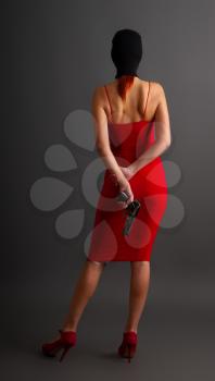 sexy girl in a tight red dress and balaclava holds an automatic pistol on her back against a dark background