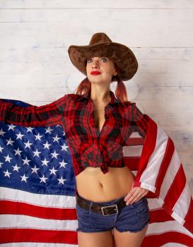 upright patriotic girl in a cowboy costume wearing a traditional wide-brimmed hat and shertach posing against a wooden background with the flag of the USA.