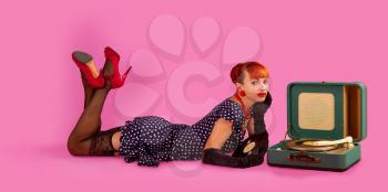 Pin-up model girl in retro polka-dot dress listens to an old gramophone on a pink background