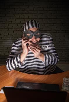 Comic character prisoner telephone con man sitting at a laptop in a striped robe and a hat in a black mask and cheating on the phone.