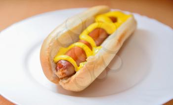 A cooked hot dog in a plain soft bun with mustard on a white plate.