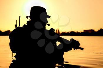 Silhouette of special forces with rifle in action during river raid in the jungle waist deep in the water. Proflie side view, half length