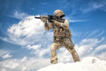 War game player in military camouflage uniform, equipped with tactical ammunition, aiming with service rifle replica while moving in sand with cloudy sky on background, during airsoft game event
