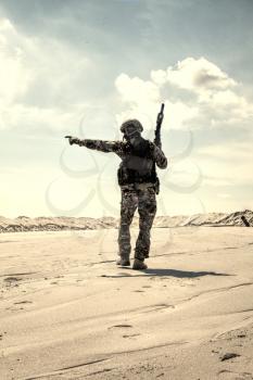 Soldier of special forces, infantry rifleman in military ammunition walking in desert and pointing on horizon. Military reconnaissance team leader managing dead ground observation with combat patrol