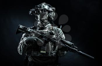 Army infantry in battle uniform, armed assault rifle with laser sight and silencer, standing in darkness, looking through night vision goggles, low key half length, studio portrait on black background